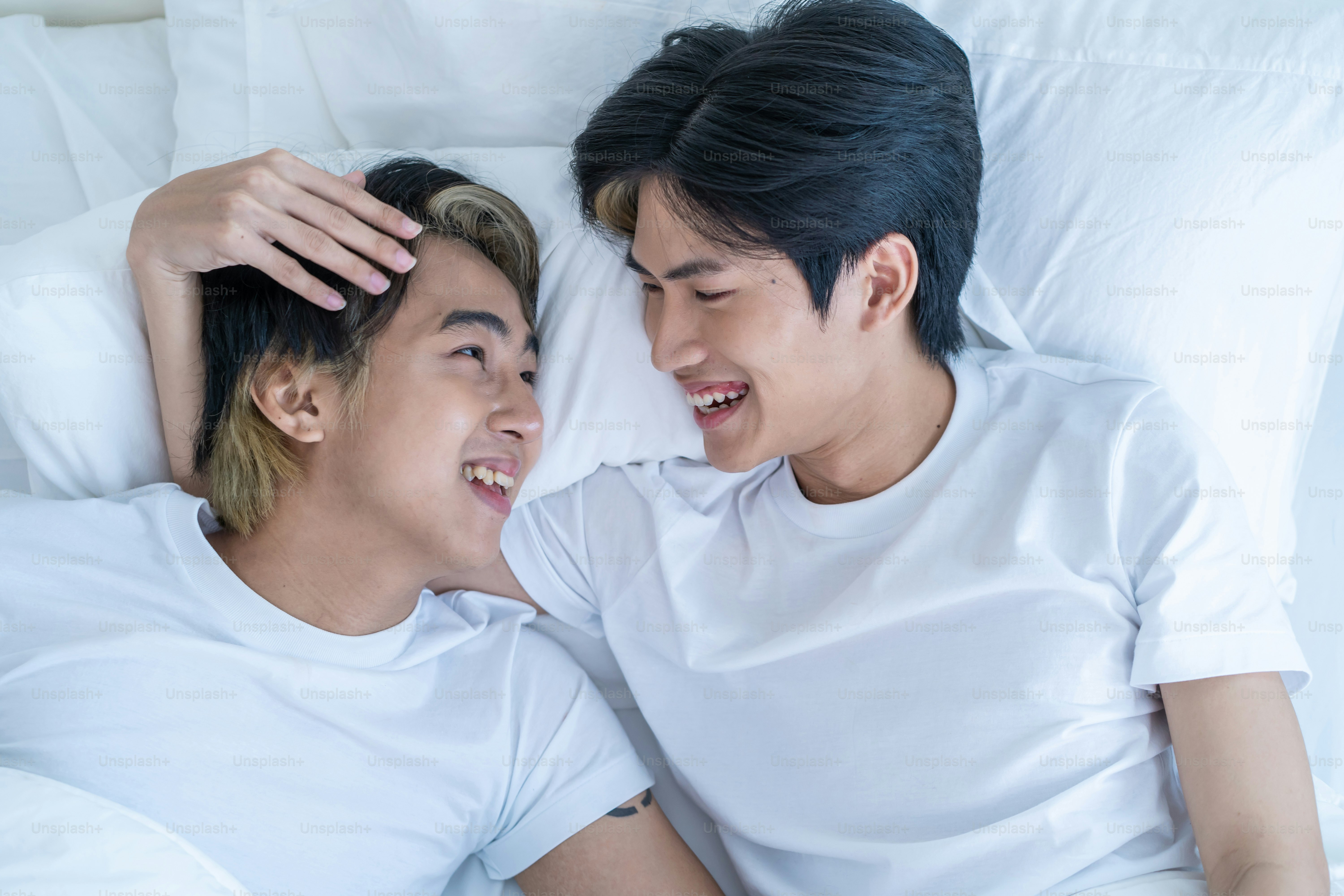 Two young men in white t-shirts lying on a bed, smiling at each other